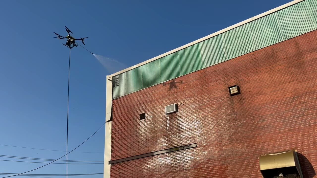 Tethered Rotag PT Services cleaning drone rinses a brick masonry wall at an industrial property.